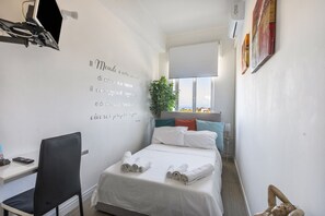 A hotel room with a bed, a desk, a chair, a potted plant, and a wall with a quote.