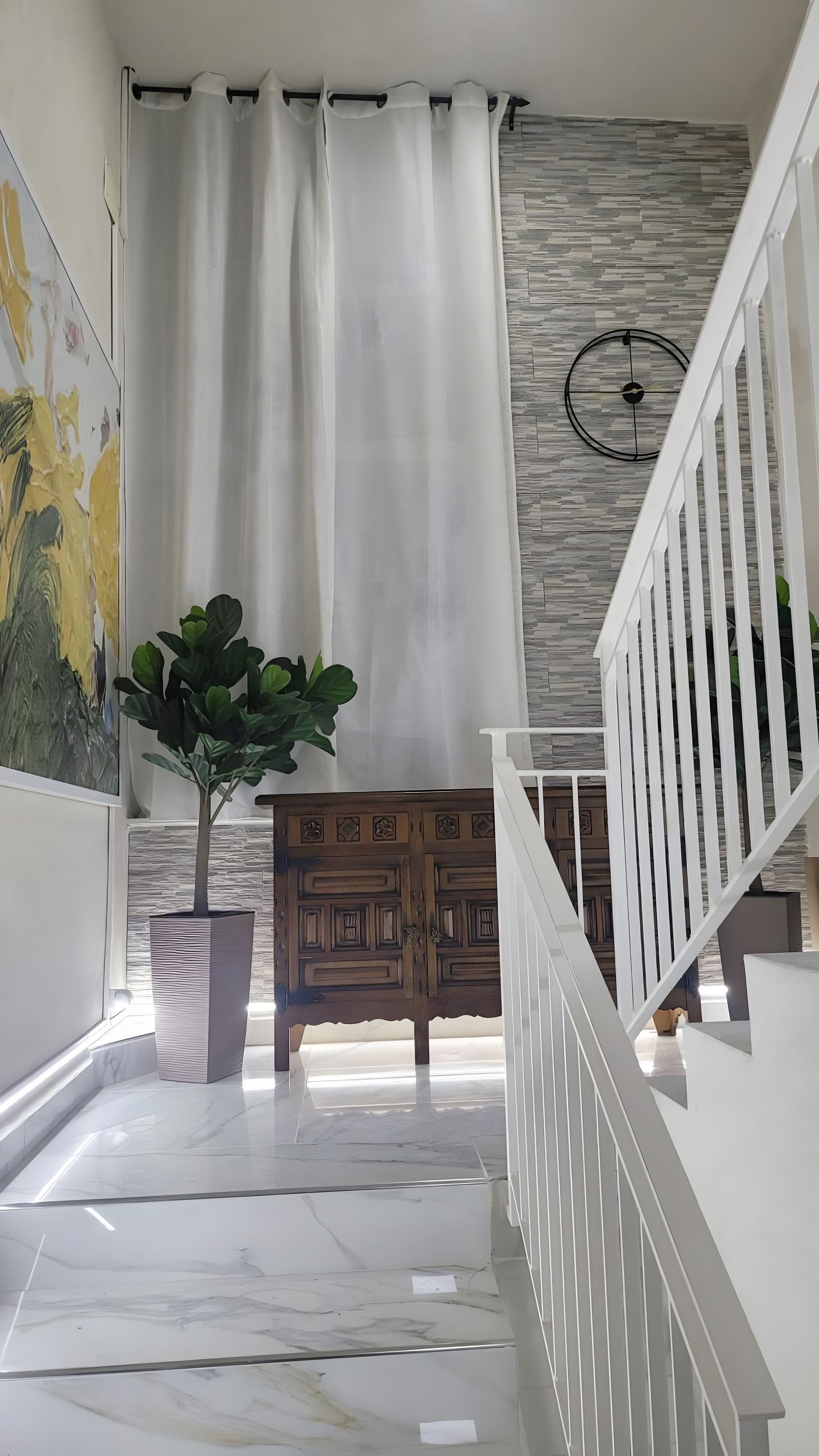 A hallway with a white staircase, a wooden cabinet, a potted plant, and a wall with a geometric pattern.