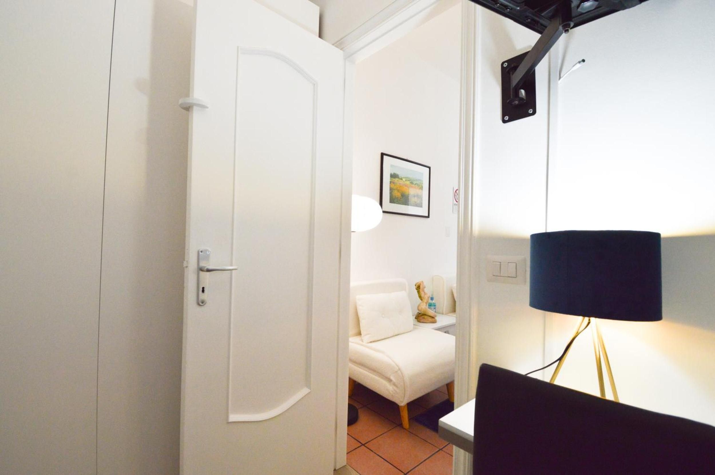 Quarto com porta branca, sofá branco, quadro na parede, luminária e escrivaninha com um computador.