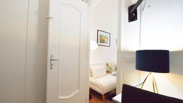 Quarto com porta branca, sofá branco, quadro na parede, luminária e escrivaninha com um computador.