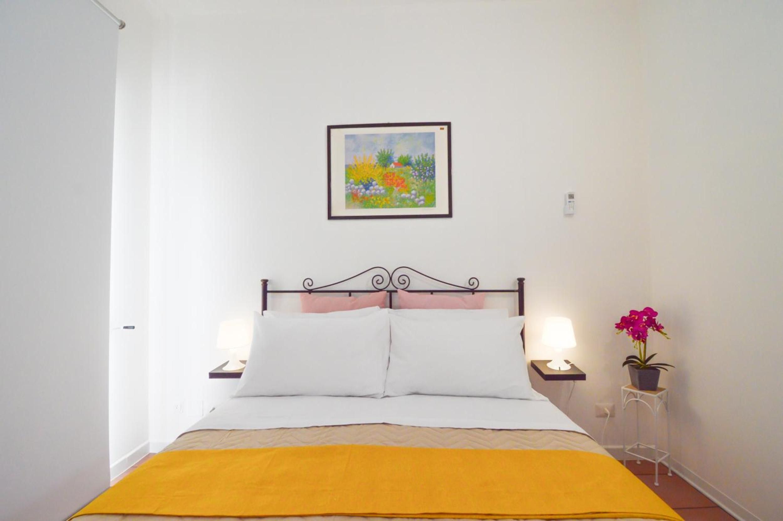 Ein Bett mit Metallkopfteil, weißer Bettwäsche und einer gelben Decke. Darüber hängt ein gerahmtes Bild und auf dem Nachttisch steht ein Blumentopf.
