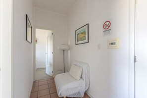 A hallway with a white chair, a framed picture, a smoke detector, and a door leading to another room.