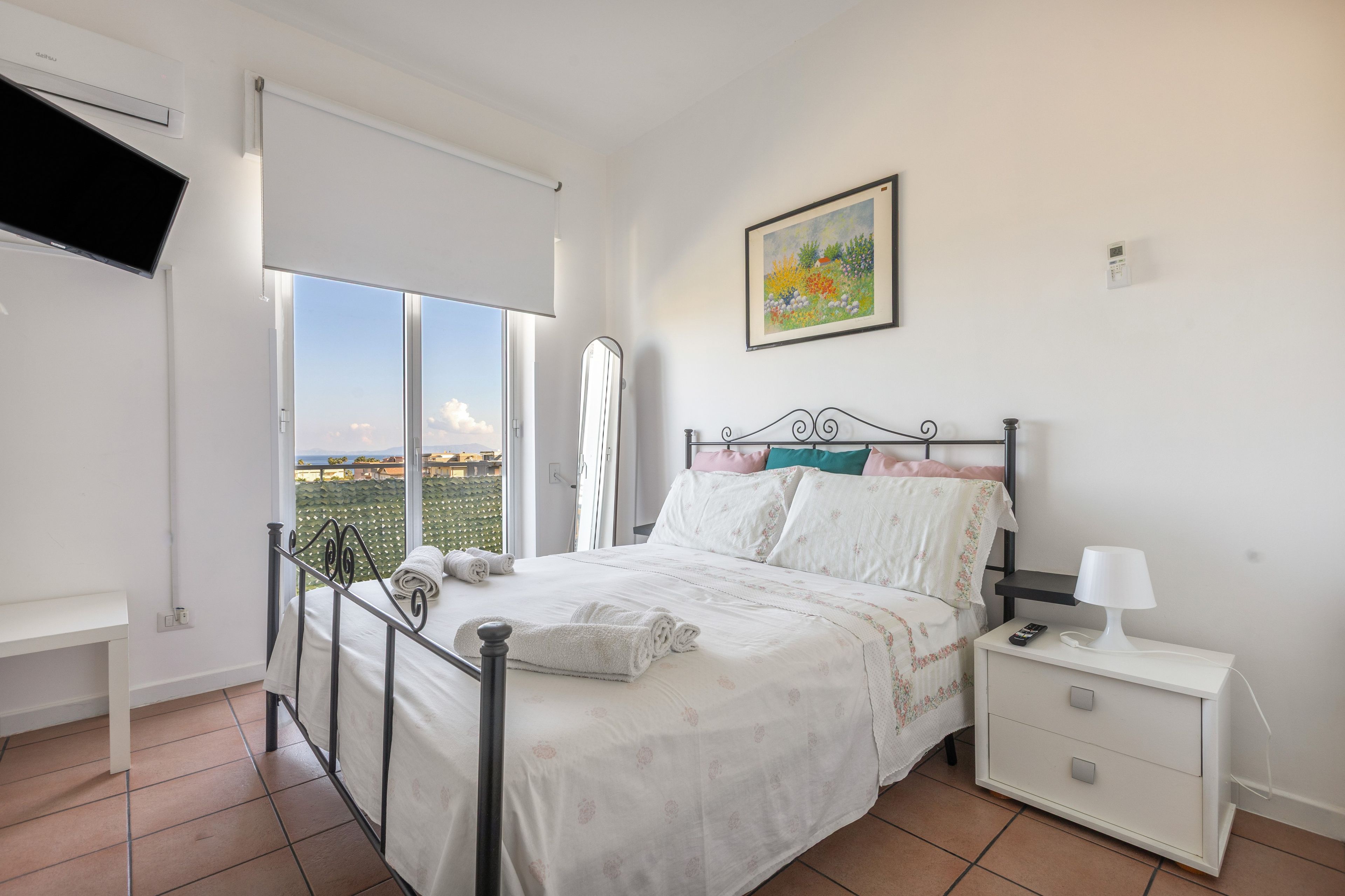 A bedroom with a bed, a nightstand, a bedside table, a window, and a painting on the wall.