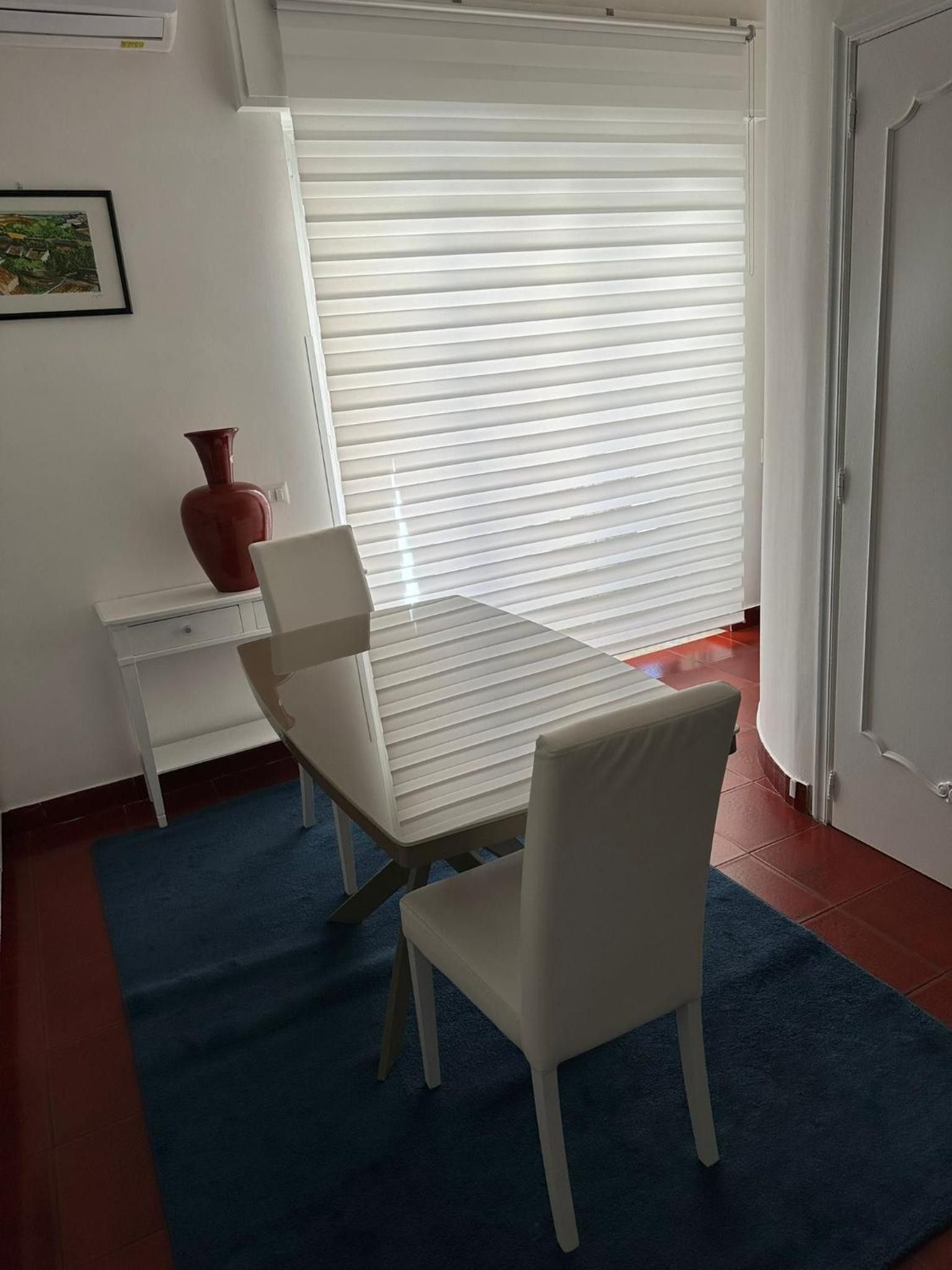 Une pièce avec une table blanche, deux chaises blanches, un vase rouge et une grande fenêtre avec des stores horizontaux.