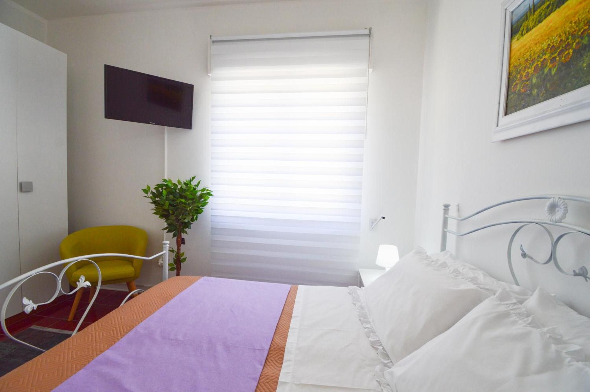 Ein Hotelzimmer mit Bett, Wandfernseher, Fenster mit Jalousien, Stuhl und einer Pflanze.