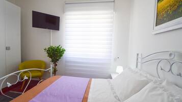 Ein Hotelzimmer mit Bett, Wandfernseher, Fenster mit Jalousien, Stuhl und einer Pflanze.