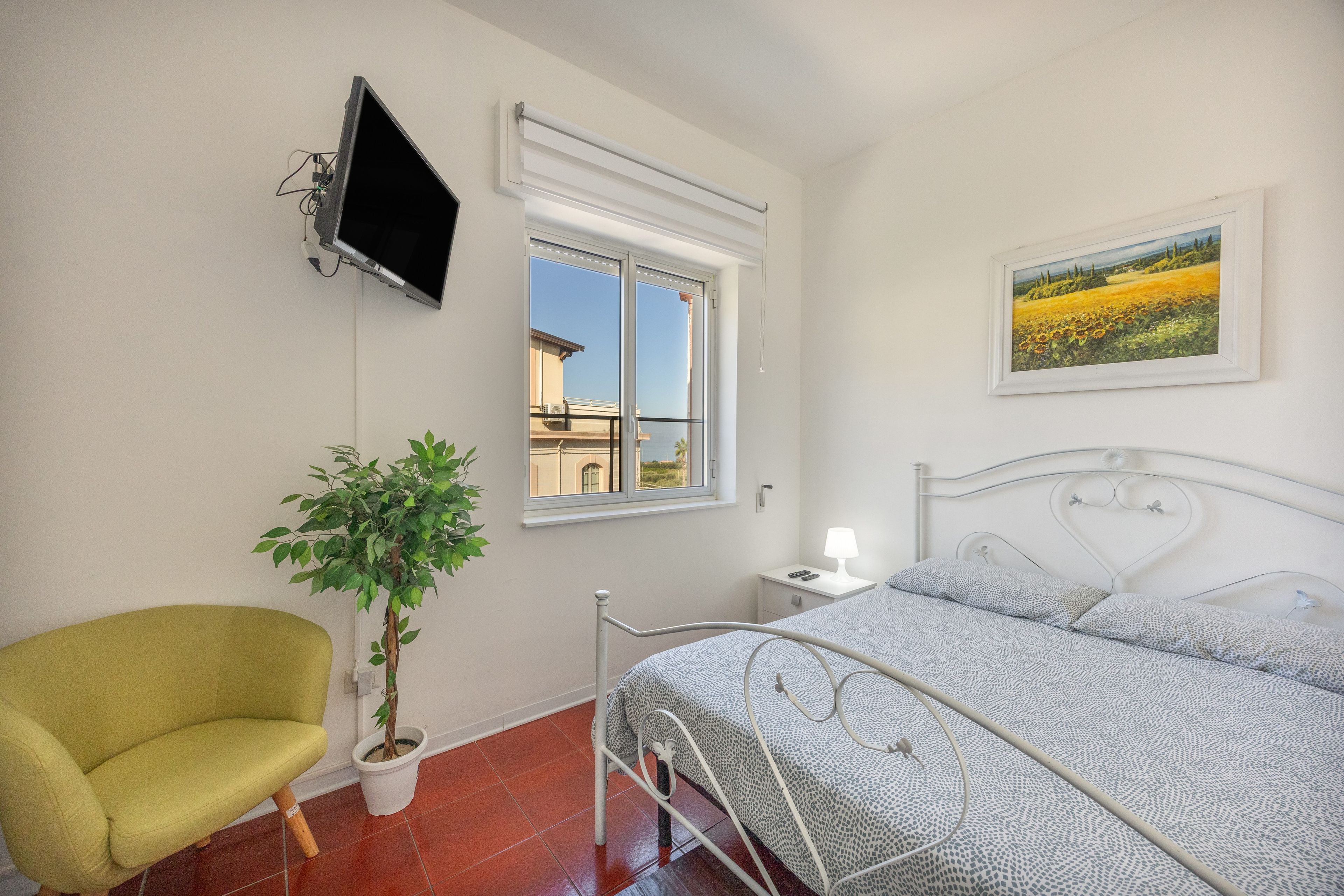 A bedroom with a bed, a chair, a potted plant, a TV, and a window with a view.