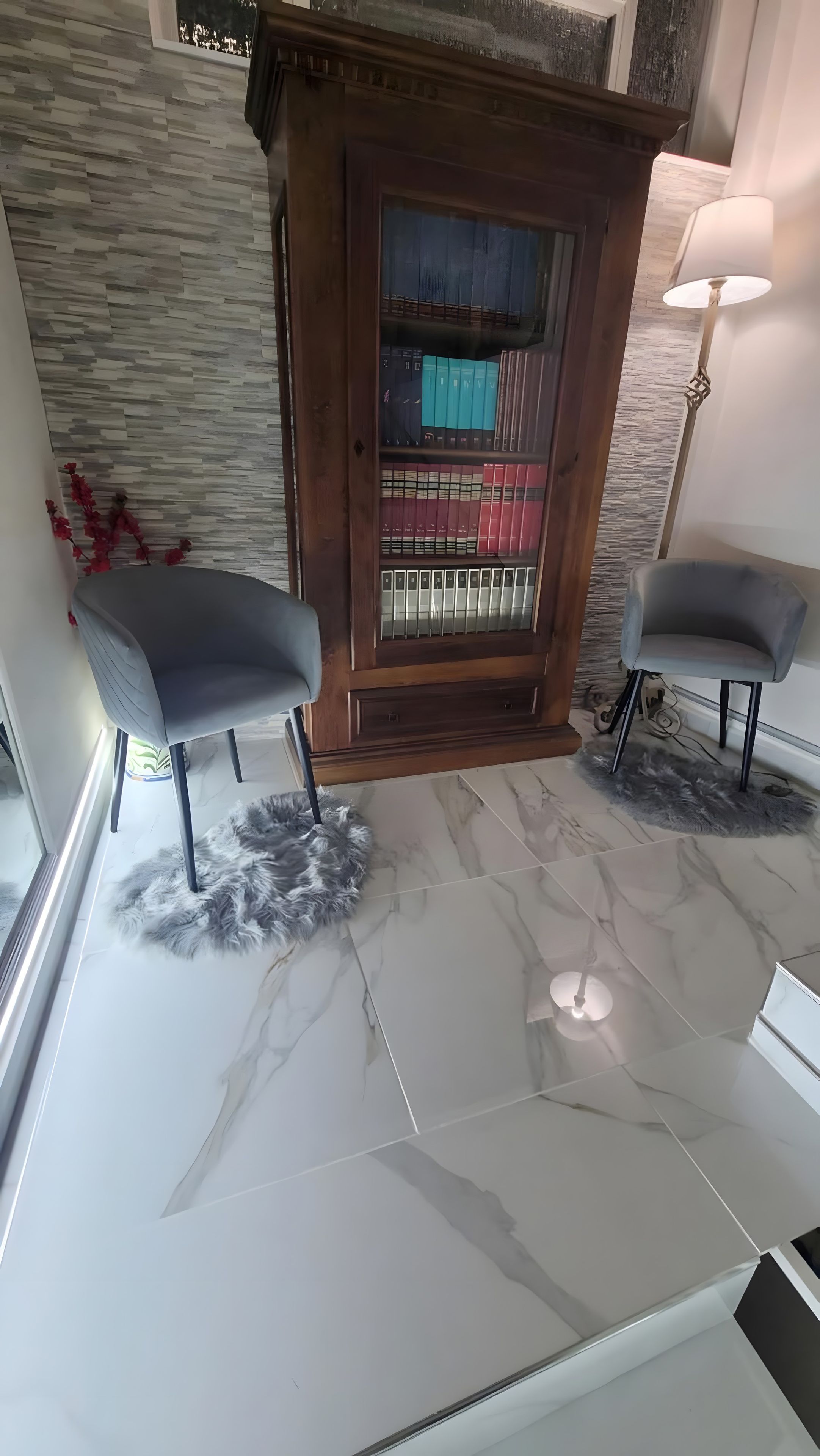 Ein modernes Interieur mit Marmorboden, zwei grau gepolsterten Sessel, einer Holztruhe mit Glastüren und einer Steinwand.