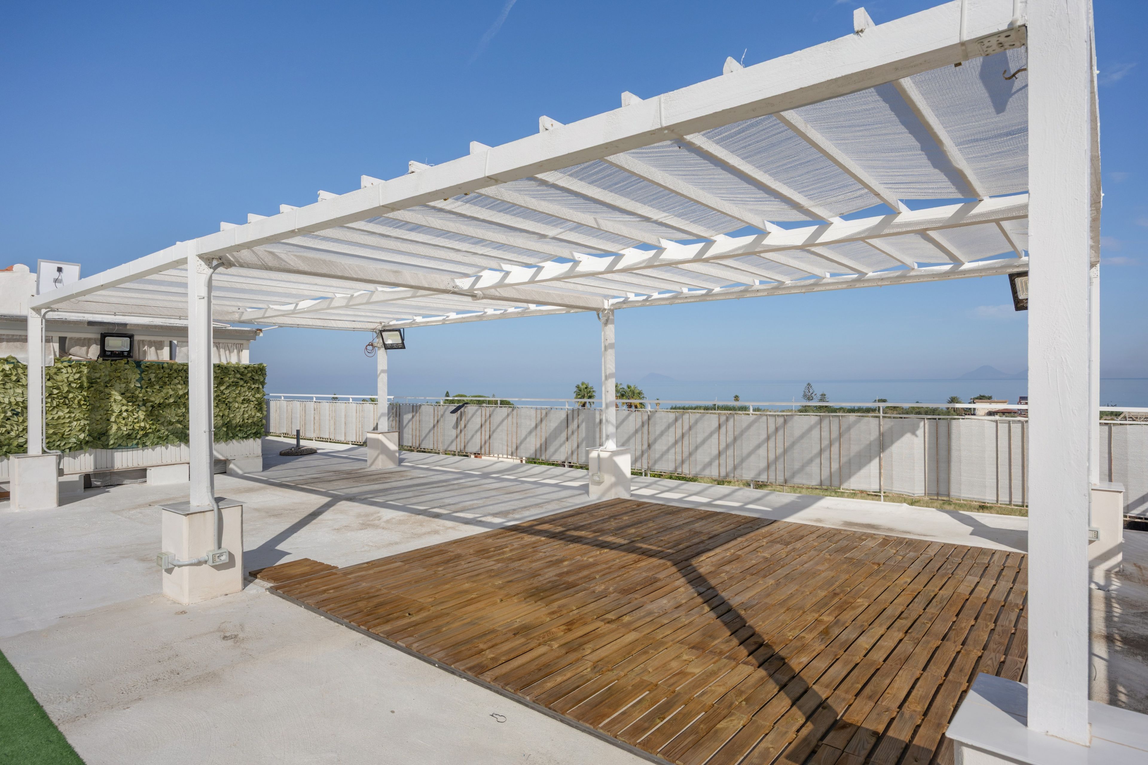 A covered outdoor deck with a wooden floor, white pergola, and a view of the sea.