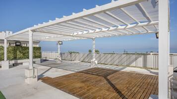 A covered outdoor deck with a wooden floor, white pergola, and a view of the sea.