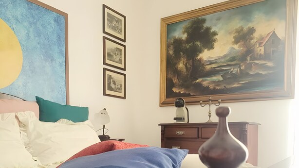Quarto com cama, travesseiros, mesa de cabeceira, luminária, cômoda e quadros na parede.