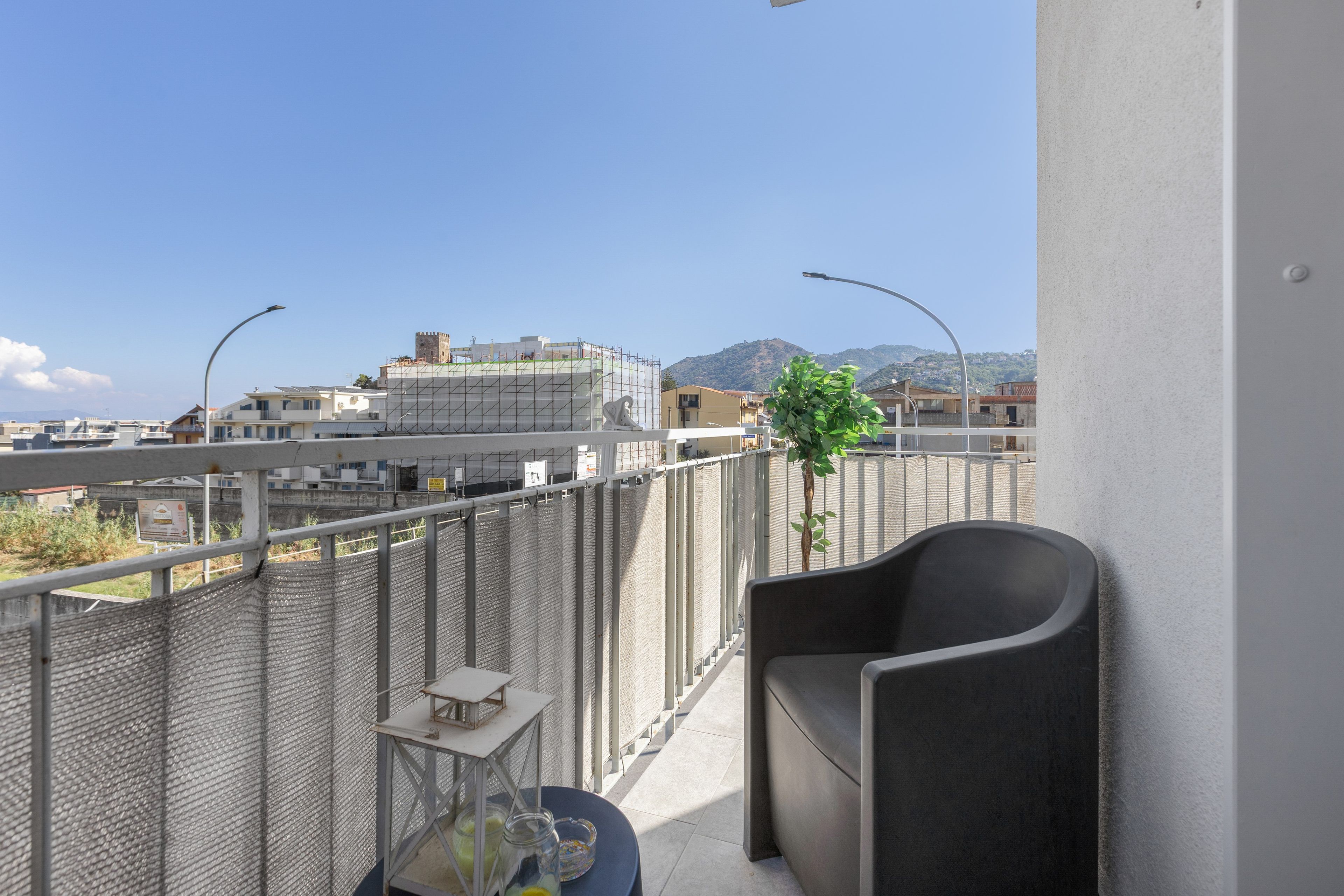 Ein Balkon mit einem Stuhl, einem kleinen Tisch und einem Topfpflanzen. Im Hintergrund sind Gebäude und ein Berg zu sehen.