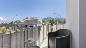A balcony with a chair, a small table, and a potted plant. In the background, there are buildings and a mountain.