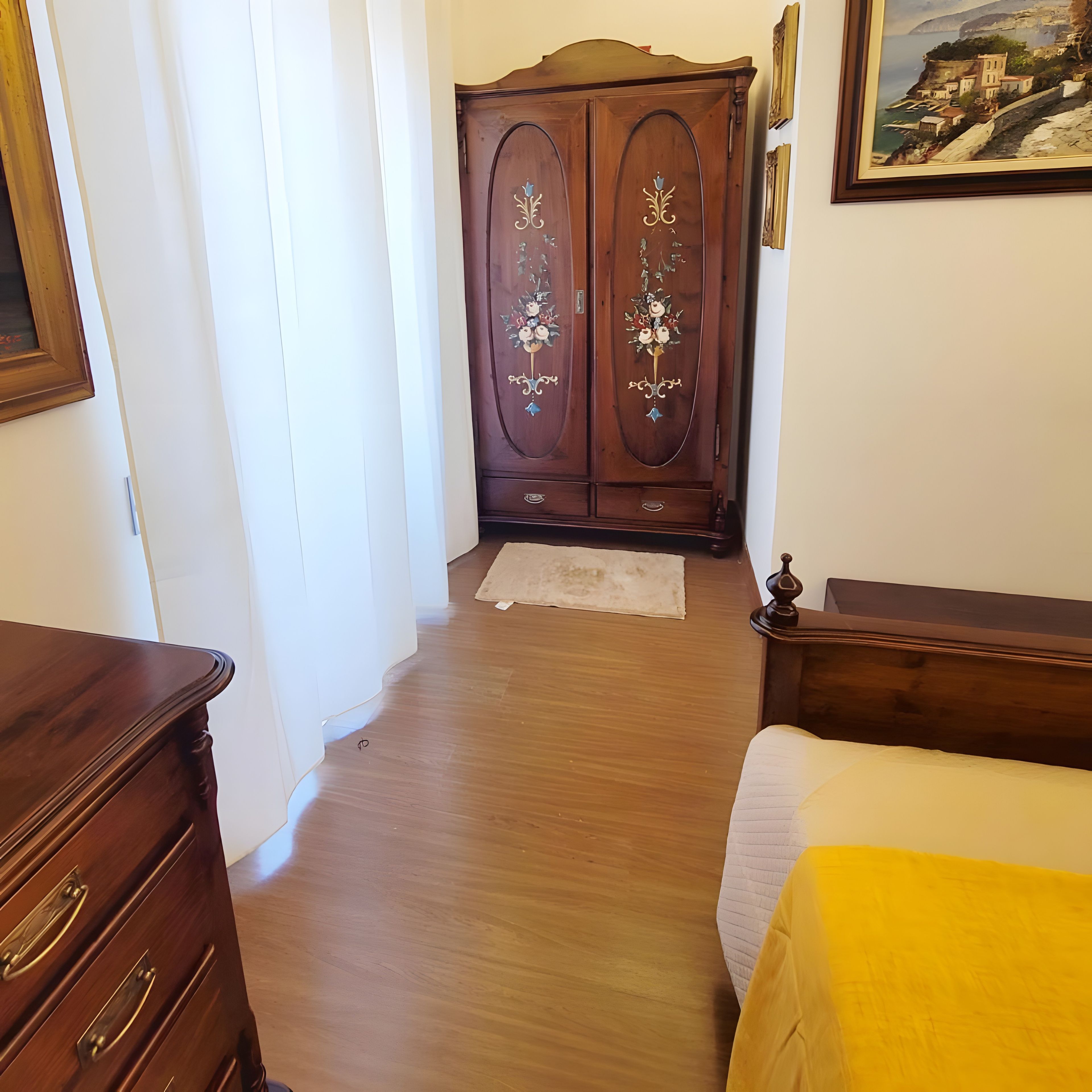 A room with a wooden wardrobe, a bed with a yellow bedspread, and a painting on the wall.
