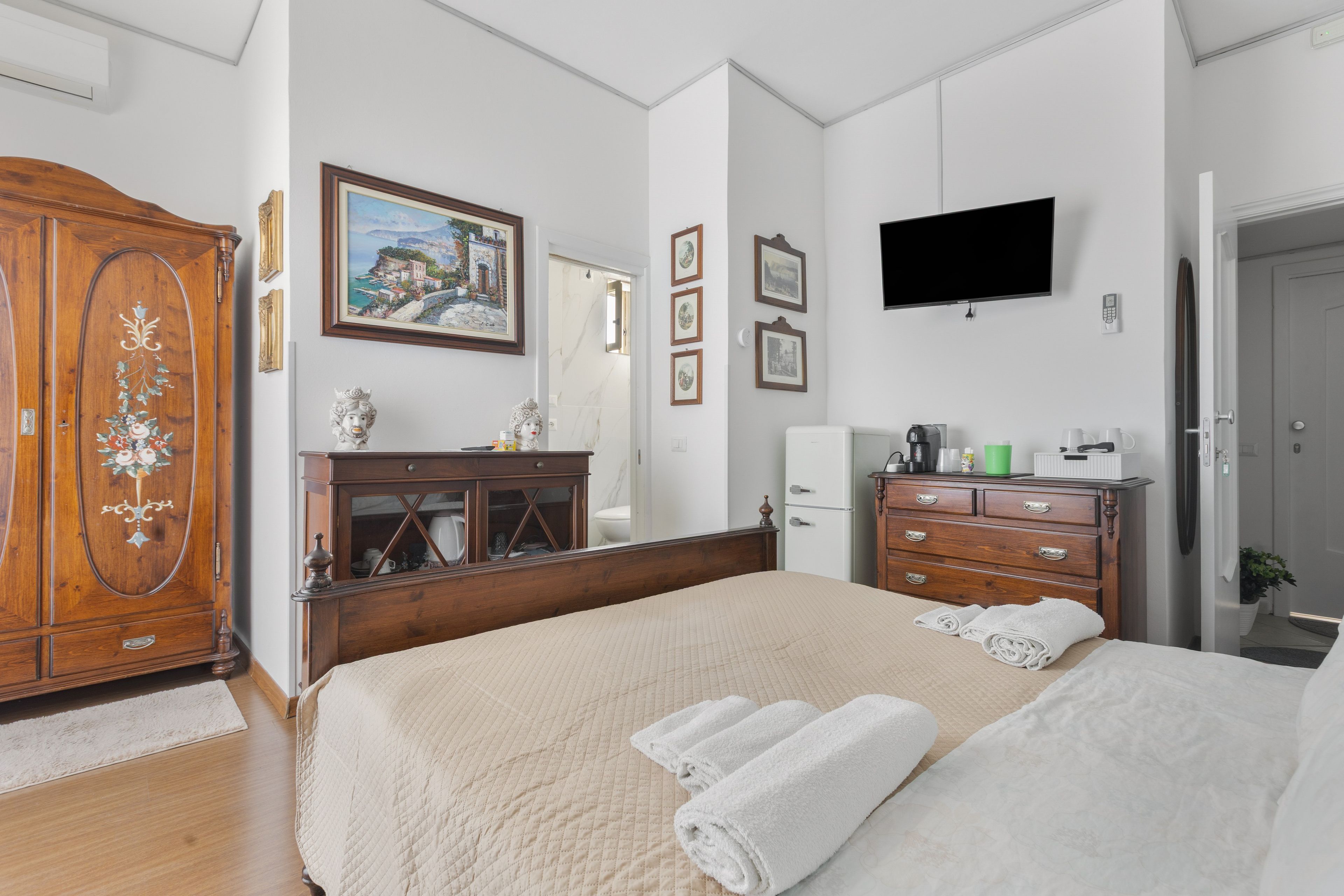 A bedroom with a bed, wooden wardrobe, dresser, and a flat-screen TV mounted on the wall.