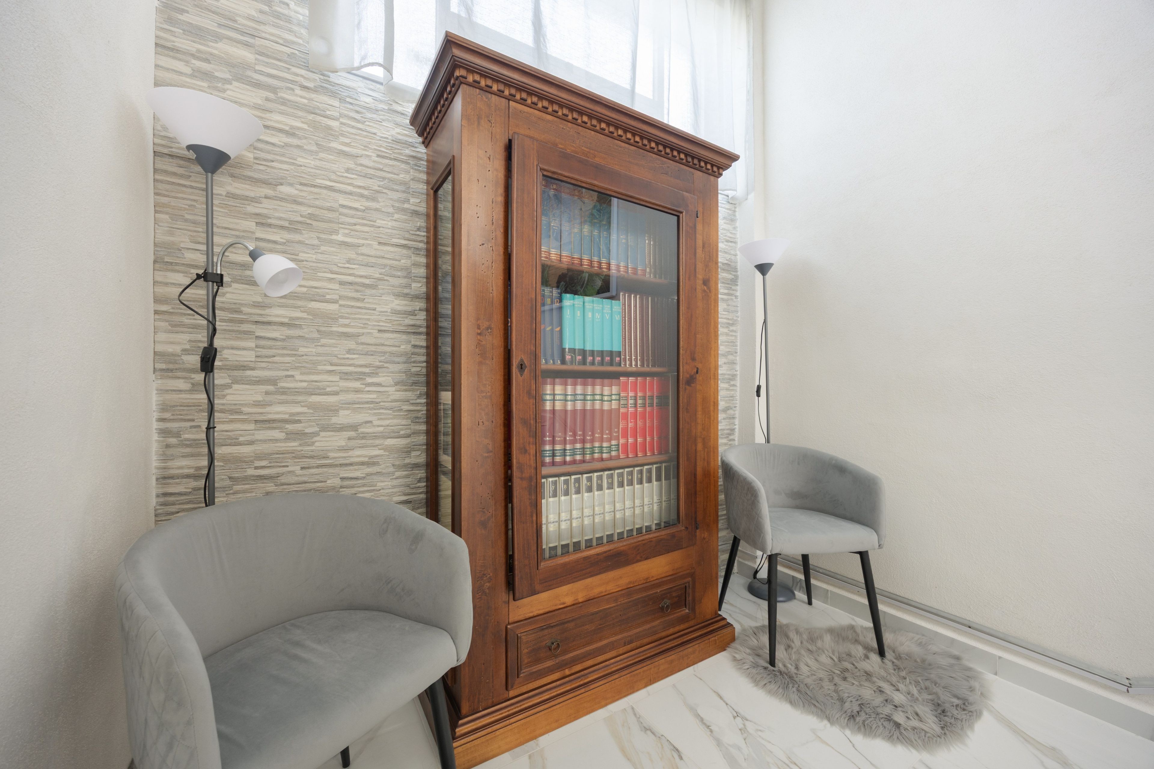 A room with a wooden bookcase, two armchairs, a marble floor, and a white rug.