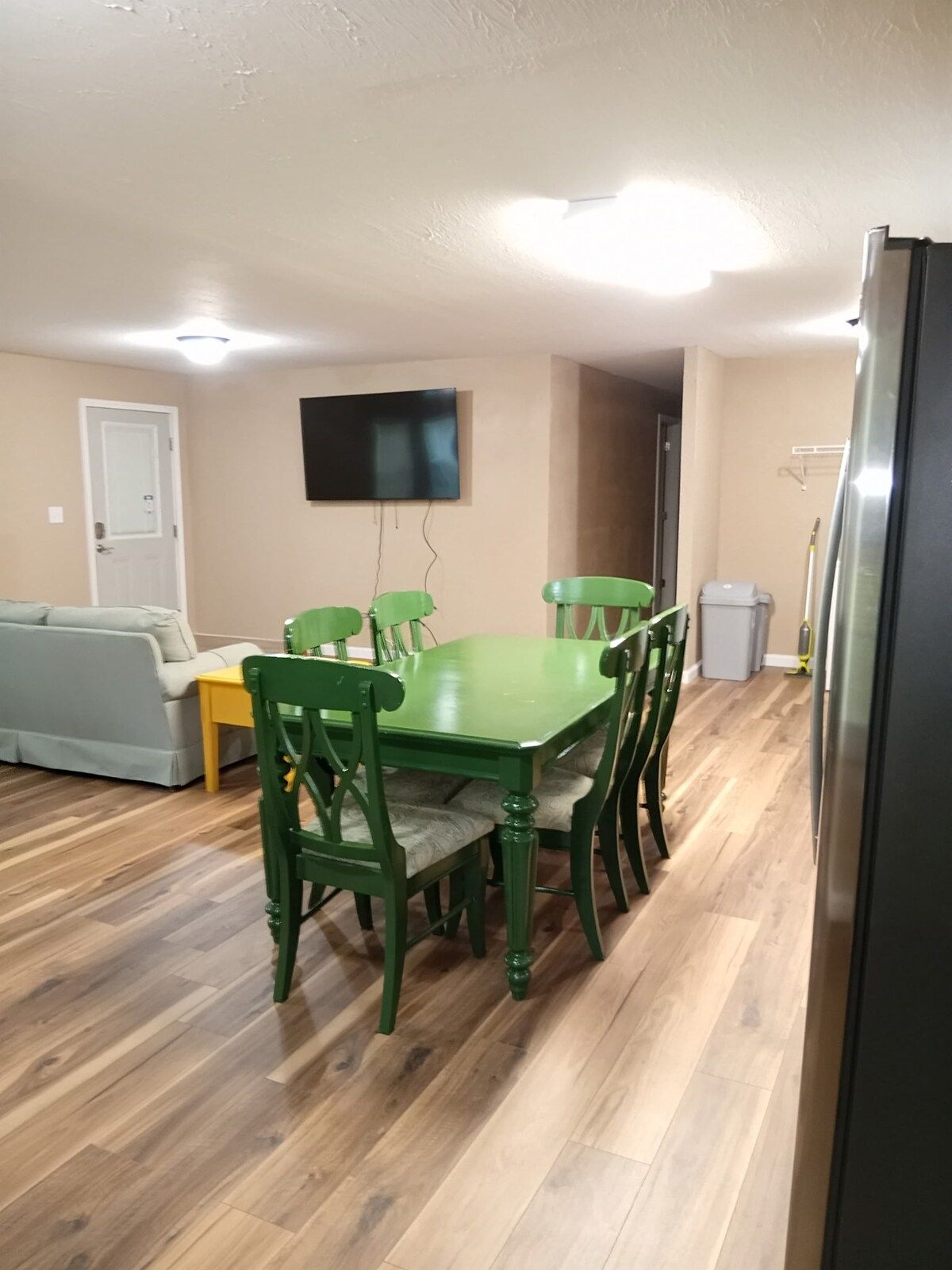 A dining area with a green table and chairs, a grey sofa, a flat-screen TV on the wall, and a door in the background.