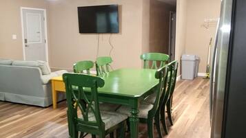 A dining area with a green table and chairs, a grey sofa, a flat-screen TV on the wall, and a door in the background.