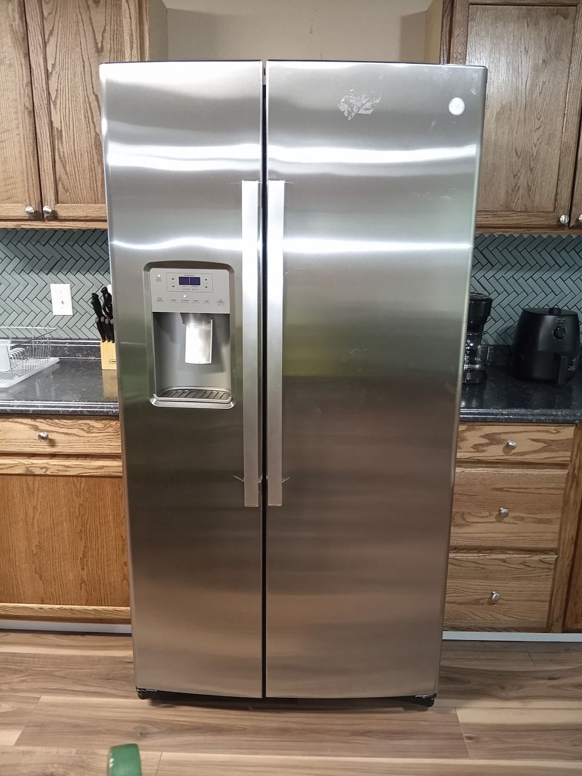 Un refrigerador de acero inoxidable con dispensador de agua, ubicado en una cocina con armarios de madera y salpicadero de baldosas tipo herringbone.
