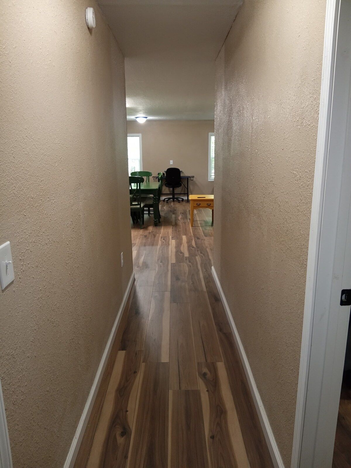 A hallway with wooden flooring and beige walls, leading to a room with a green chair, a black office chair, and a small table.