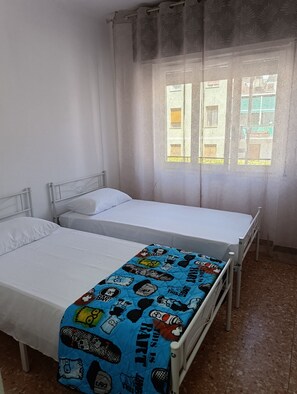 Une chambre avec deux lits, l’un recouvert d’une simple couverture blanche et l’autre d’une couverture colorée à motifs de Mickey Mouse. Il y a une fenêtre avec des rideaux, et la chambre est dotée d’un sol carrelé.