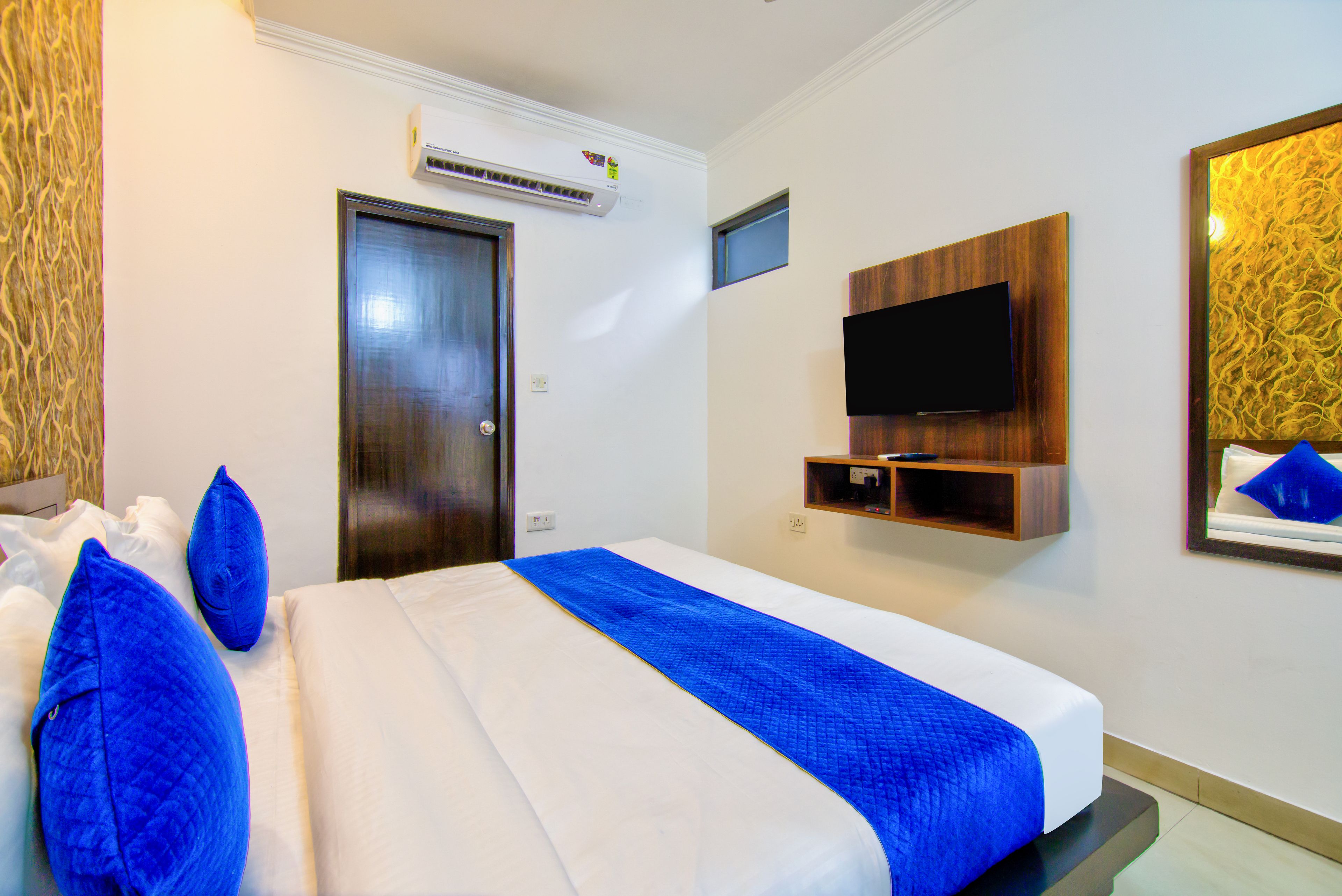Una habitación de hotel con una cama, un televisor montado en la pared, un equipo de aire acondicionado sobre la puerta y un estante pequeño debajo del televisor.