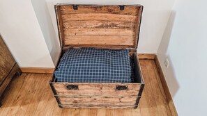 Ein offener Holzkoffer mit blau kariertem Futter, auf einem Holzboden vor einer weißen Wand platziert.