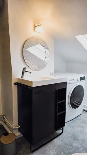 Een moderne badkamer met een witte wastafel, een ronde spiegel, een zwart kastje en een voorlader wasmachine.