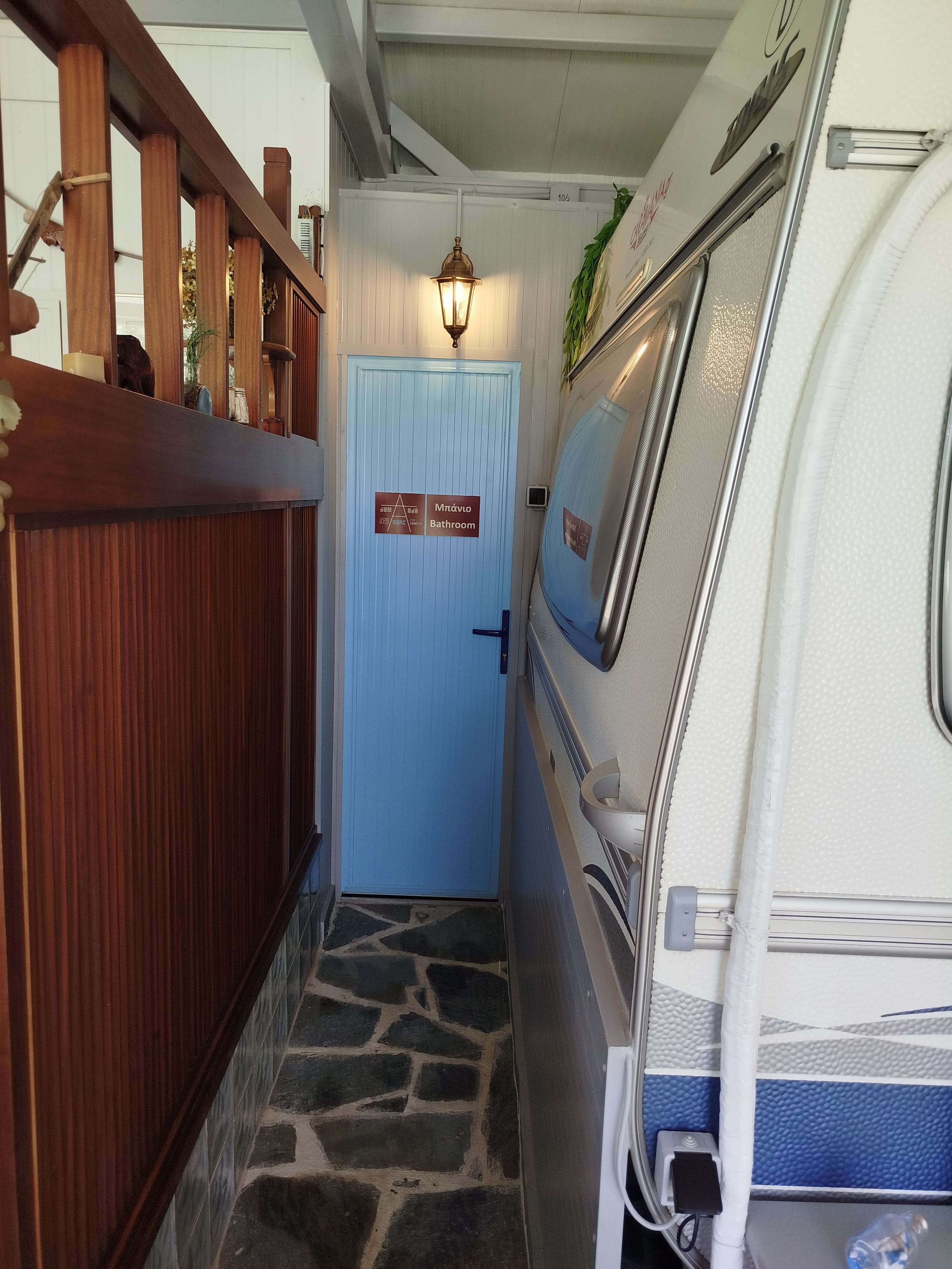 A narrow hallway with a blue door labeled 'Mr. Ávila Bathroom,' a wooden railing on the left, and a white RV parked on the right.