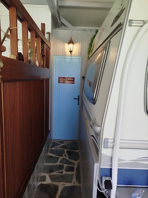 A narrow hallway with a blue door labeled 'Mr. Ávila Bathroom,' a wooden railing on the left, and a white RV parked on the right.