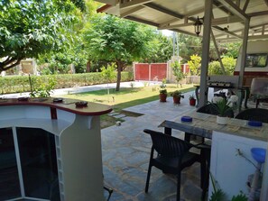 Een terras buiten met een tafel en stoelen, omringd door planten in potten en uitzicht op een zwembad en tuin.
