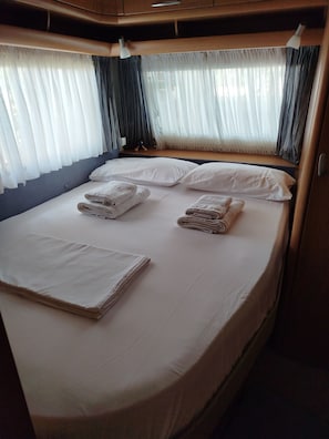 Een net opgemaakt bed met kussens en opgevouwen handdoeken, een raam met dunne gordijnen en een plafondlamp.