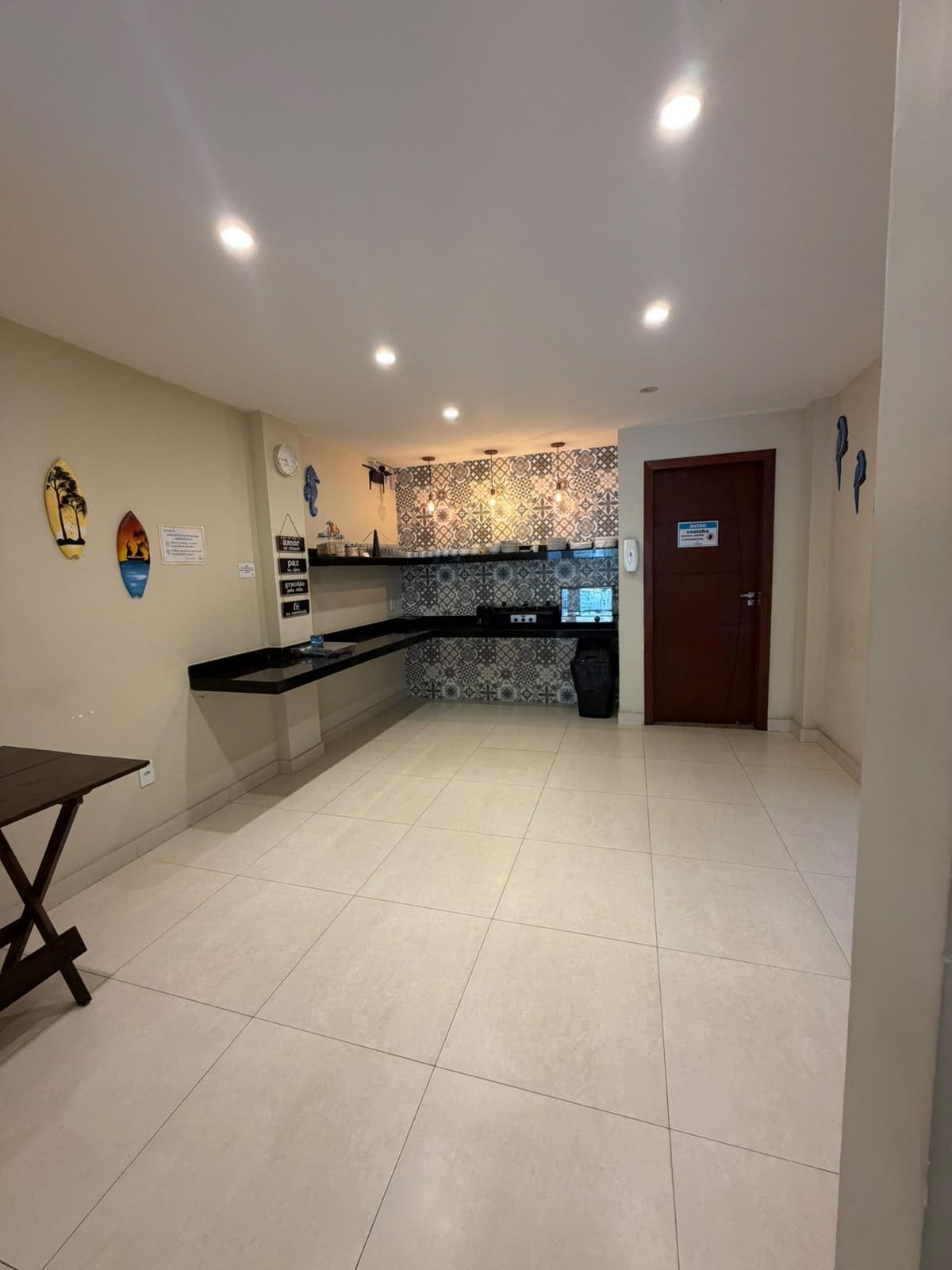 A spacious kitchen with a tiled floor, a black countertop, and a patterned backsplash. There is a wooden door and a wall-mounted clock.