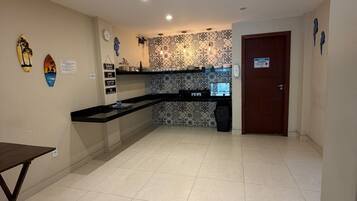 A spacious kitchen with a tiled floor, a black countertop, and a patterned backsplash. There is a wooden door and a wall-mounted clock.