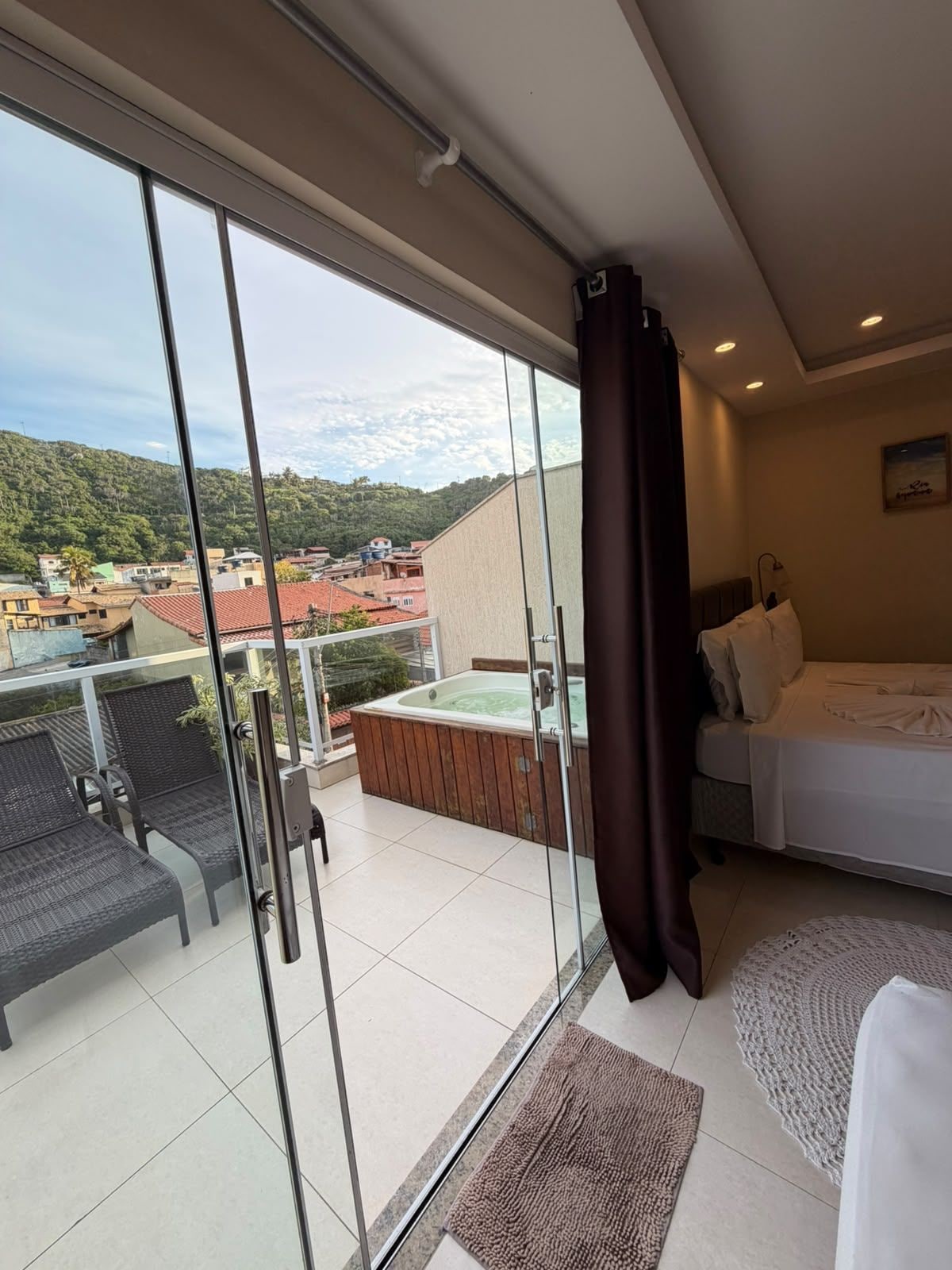 A balcony with a hot tub, a bed, and a view of buildings and hills.