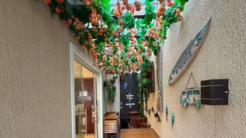 A narrow outdoor corridor with a wooden table and chairs, a door leading to a room, and a wall with a sign and decorative items.