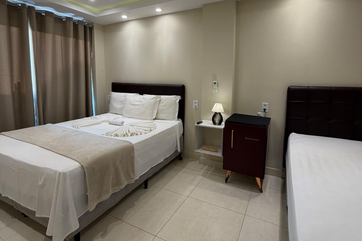 A hotel room with a double bed, a bedside table with a lamp, a small cabinet, and a window with curtains.