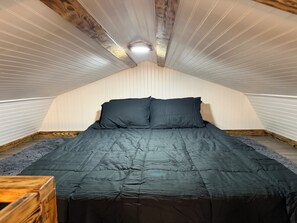 A room with a sloped ceiling, wooden beams, a bed with dark bedding, and a small wooden table. - The Marie: Cozy East Texas Stay Near Great Fishing (Quitman)