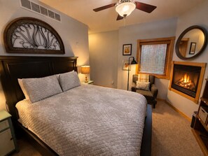 A bedroom with a bed, bedside lamps, a chair, a fireplace, and a ceiling fan. - Emerald Mountain 28C | Couples Retreat at Mary’s Lake (Estes Park)
