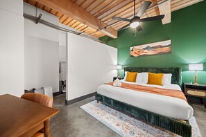 A hotel room with a bed, a desk, a chair, a ceiling fan, and a painting on the wall.