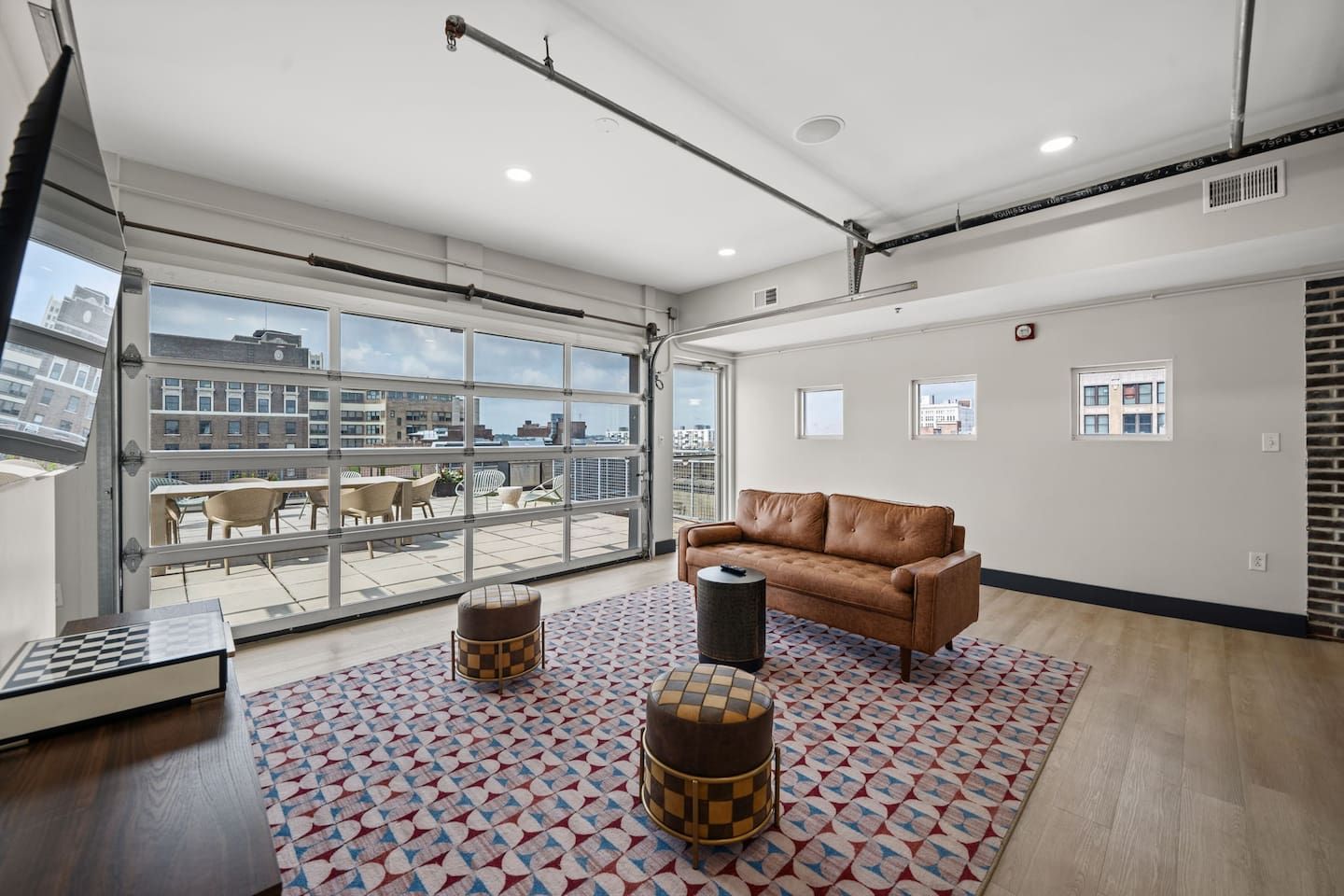 A spacious room with a large window, a brown leather sofa, a patterned rug, and a small round ottoman.