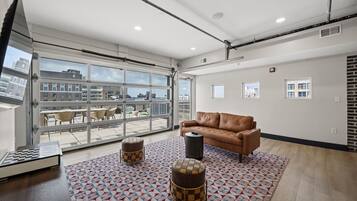 A spacious room with a large window, a brown leather sofa, a patterned rug, and a small round ottoman.