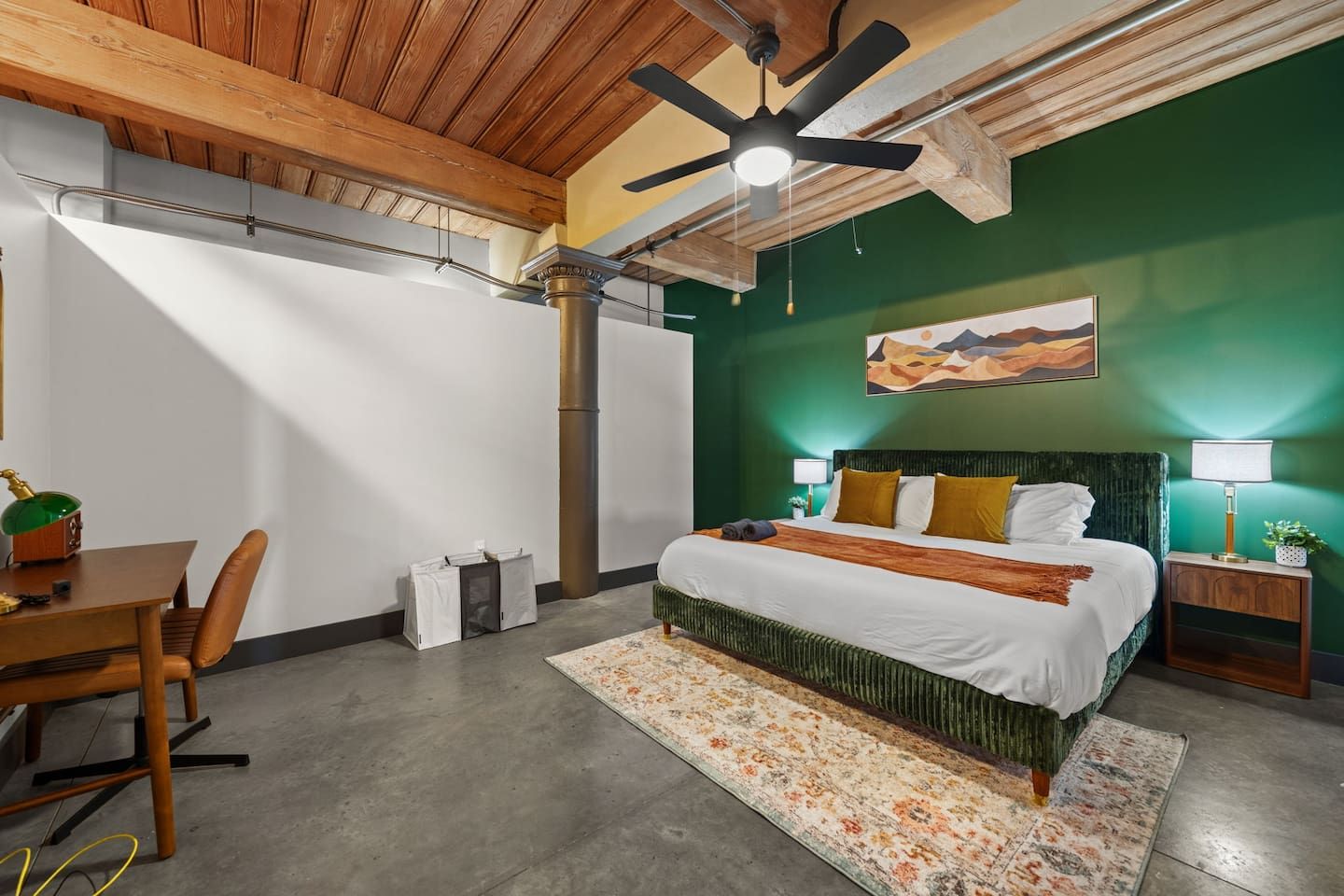 A bedroom with a bed, a desk, a chair, a ceiling fan, and a painting on the wall.