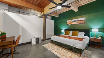 A bedroom with a bed, a desk, a chair, a ceiling fan, and a painting on the wall.