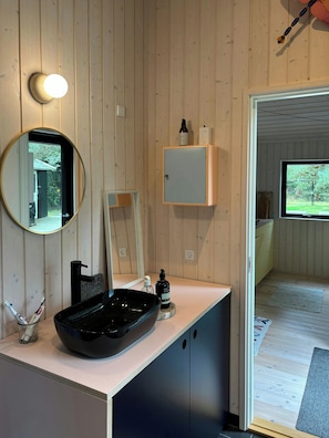 A bathroom with a wooden wall, a round mirror, a black sink, and a cabinet with toiletries. - Modern holiday home with vaulted ceiling, Tisvilde (Frederiksværk)