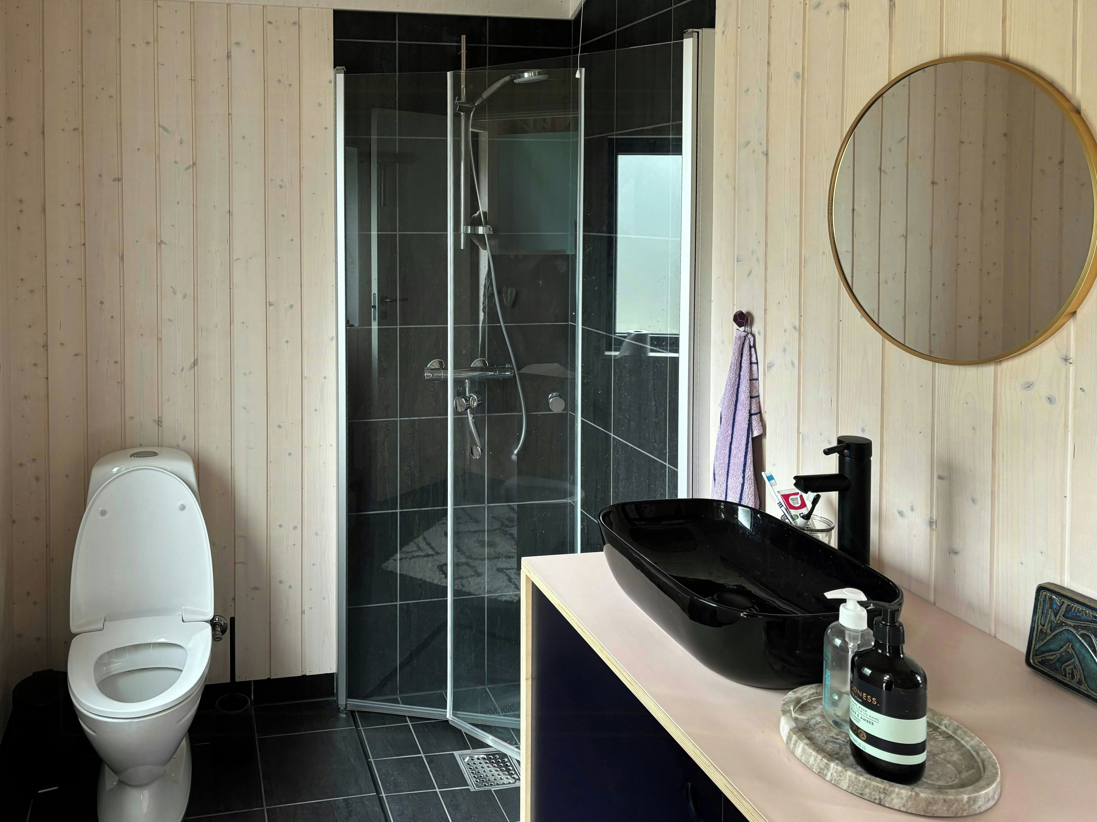 Ein modernes Badezimmer mit Toilette, einer gläsernen Duschkabine, einem runden Spiegel und einem Waschtisch mit schwarzem Waschbecken und Seifenspender.
