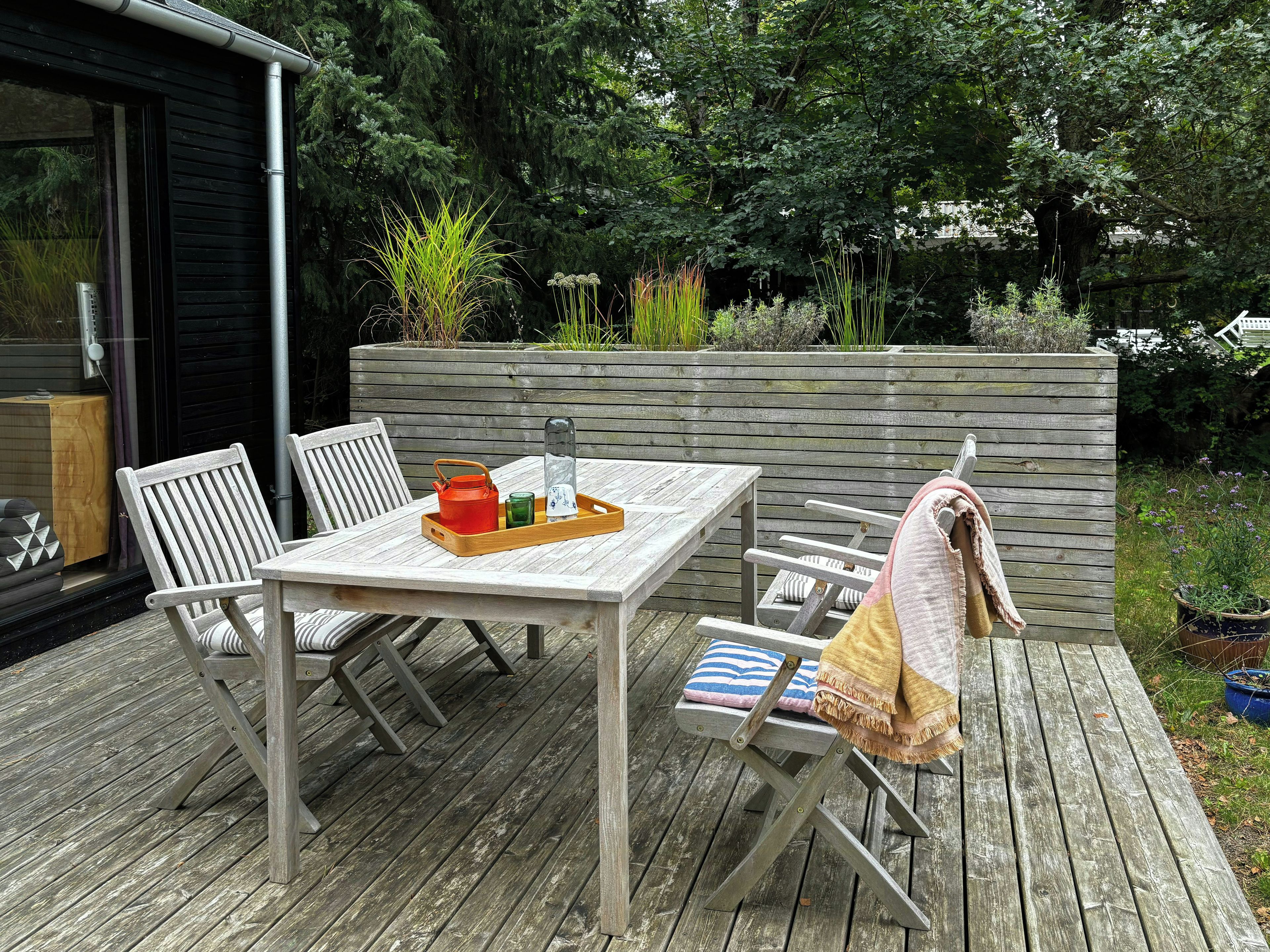 Une terrasse extérieure en bois, avec une table et des chaises, un banc avec une serviette, et un bac à fleurs en bois contenant des plantes.