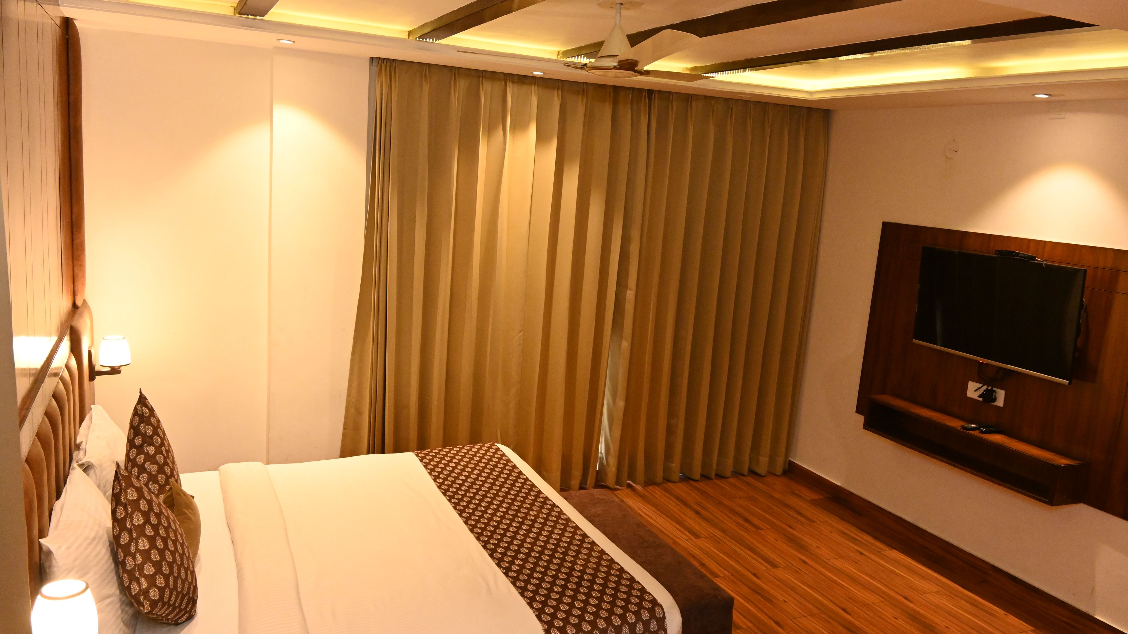 A hotel room with a bed, curtains, a television mounted on the wall, and a wooden floor.