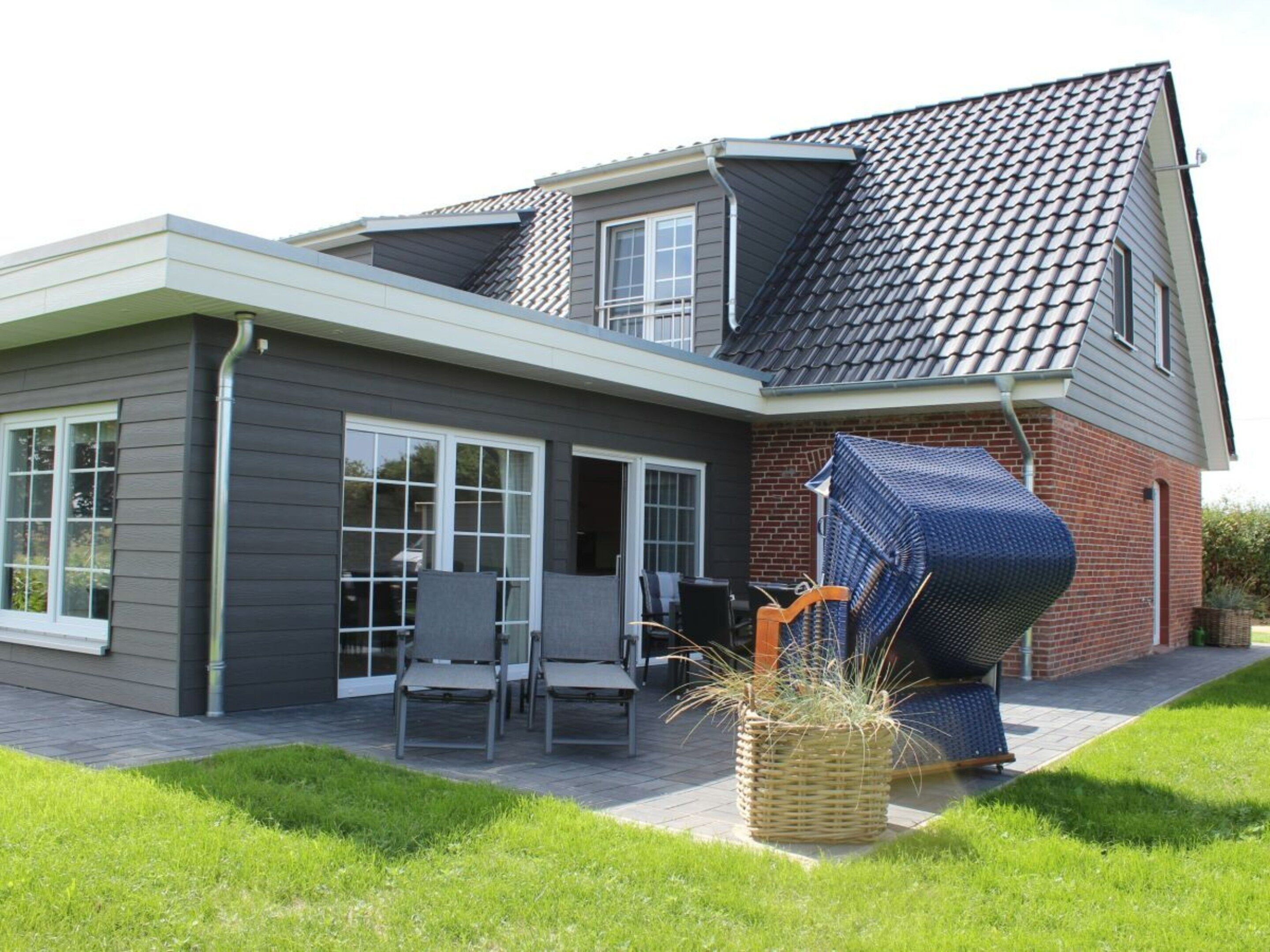 A modern house with a grey exterior, a sloped roof, and a brick section. There is a patio with outdoor furniture and a large woven basket.