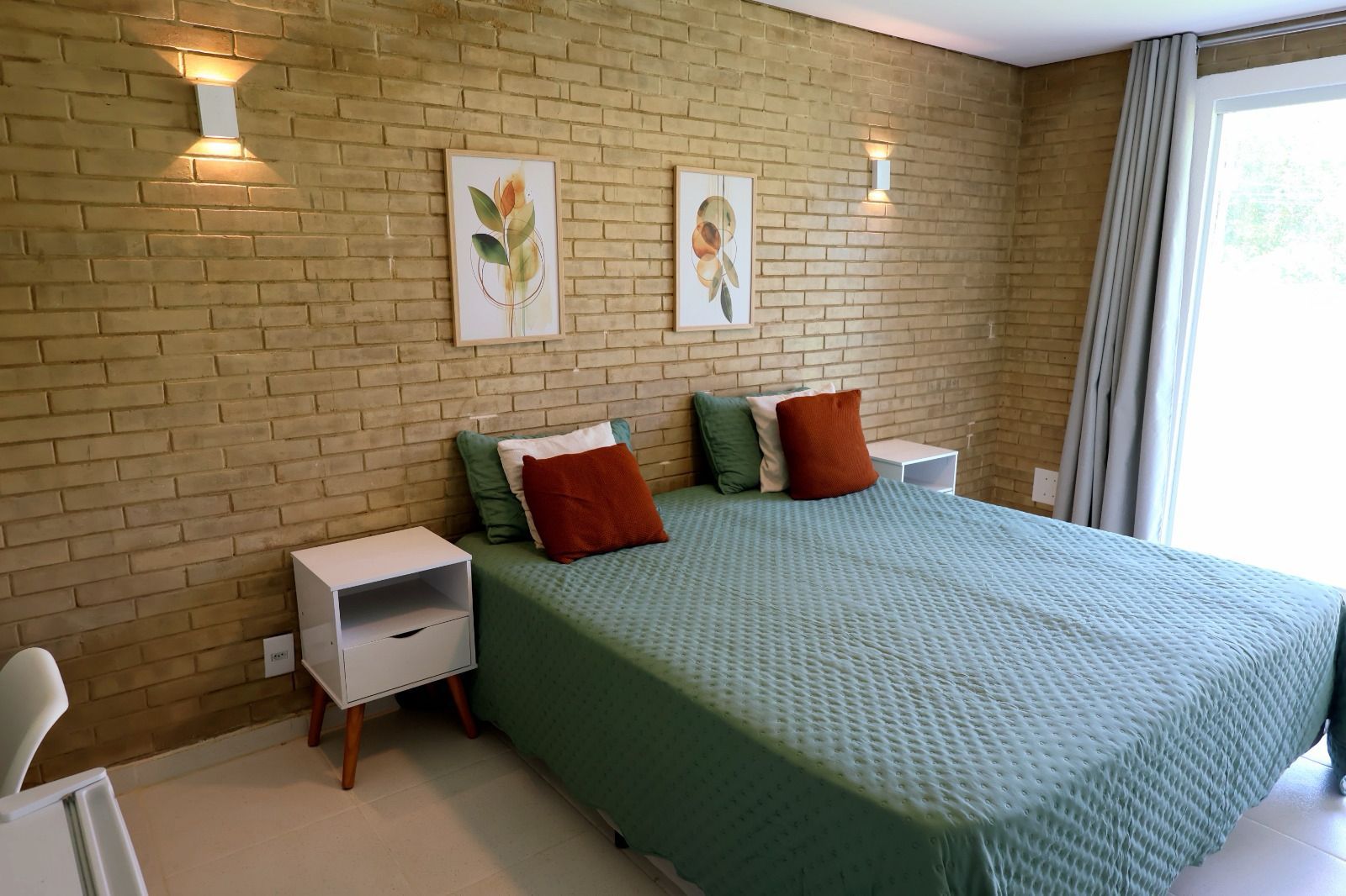 A bedroom with a brick wall, a bed with green bedding, two pillows, and two wall-mounted light fixtures.